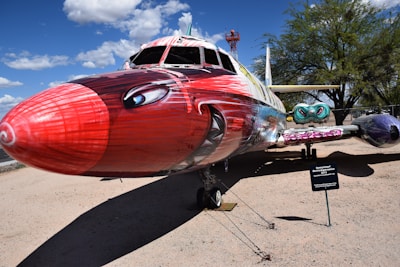 A creatively painted airplane displays vivid and colorful artwork. The nose is designed with bold red tones, adorned with eyes and other intricate patterns. The fuselage features a mix of colors, including blues and purples, with whimsical designs and faces. In the background, there are clear skies with fluffy clouds and some trees.