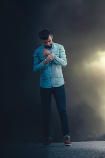 A man stands outdoors on a dark background, wearing a denim shirt and jeans, adjusting his sleeve. He appears to be deep in thought or contemplation.