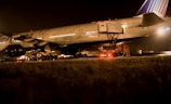 An airplane is parked on a tarmac at night under bright lights. The aircraft is being serviced with a cargo loader attached to the open door near the rear. The scene is dimly lit with a warm glow, and there are vehicles and equipment visible around the aircraft.