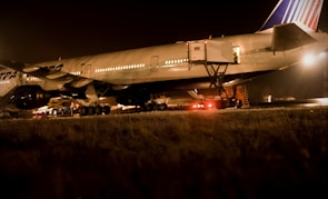 An airplane is parked on a tarmac at night under bright lights. The aircraft is being serviced with a cargo loader attached to the open door near the rear. The scene is dimly lit with a warm glow, and there are vehicles and equipment visible around the aircraft.