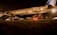 An airplane is parked on a tarmac at night under bright lights. The aircraft is being serviced with a cargo loader attached to the open door near the rear. The scene is dimly lit with a warm glow, and there are vehicles and equipment visible around the aircraft.
