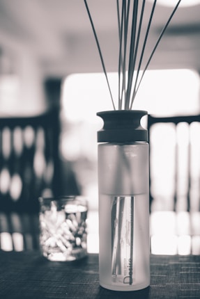 A monochromatic scene featuring a reed diffuser placed centrally on a table. The background is softly blurred, with hints of furniture creating a homely atmosphere. The diffuser bottle is translucent with visible liquid inside, topped with a dark cap and several thin reeds extending upwards. Nearby, a glass candle holder adds subtle texture to the composition.