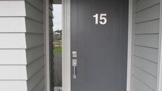 A dark gray front door with the number 15 clearly displayed in white. The door features a modern metal handle and lock. It is part of an exterior entryway with white and light gray panel siding. The view includes a glimpse outside showing a gravel path and a fence.