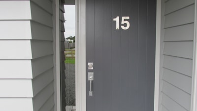 A dark gray front door with the number 15 clearly displayed in white. The door features a modern metal handle and lock. It is part of an exterior entryway with white and light gray panel siding. The view includes a glimpse outside showing a gravel path and a fence.