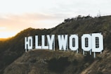 Large white letters spelling 'HOLLYWOOD' are mounted on a hillside. The landscape includes brown rocky terrain and sparse vegetation. The setting sun casts a warm glow over the scene, creating a dramatic contrast with the clear sky.