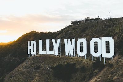 Large white letters spelling 'HOLLYWOOD' are mounted on a hillside. The landscape includes brown rocky terrain and sparse vegetation. The setting sun casts a warm glow over the scene, creating a dramatic contrast with the clear sky.