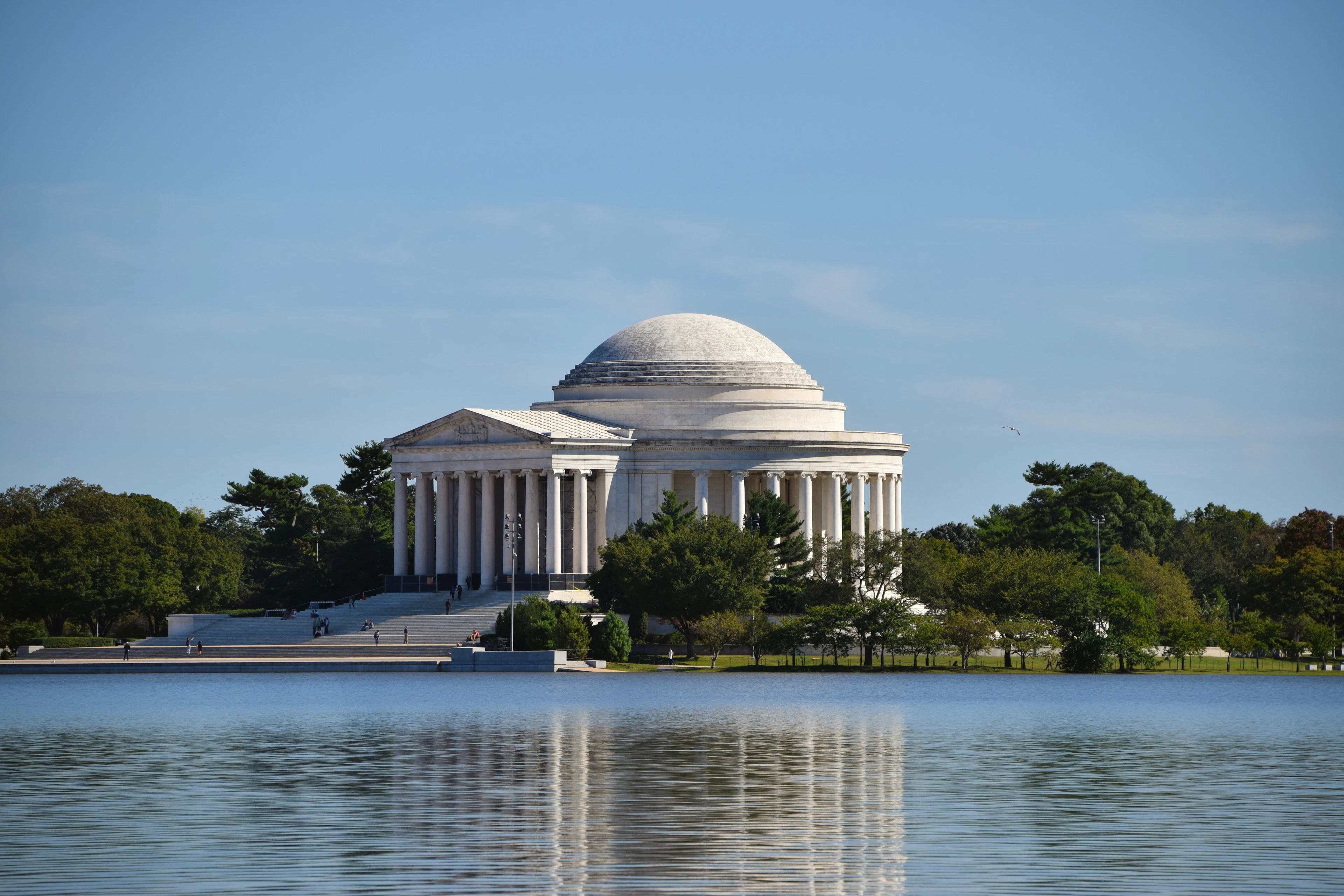 The domed Jefferson Memorial standing in its ring of trees across the Tidal Basin in Washington, D.C., its white marble colonnade mirrored in the still water under a clear blue sky