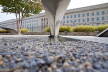 A stone and gravel ground leads to a monument-like structure with angular metal elements. A large institutional building with numerous windows stands in the background, bordered by a fence. Greenery such as trees and grass are present around the structure.