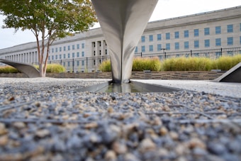 A stone and gravel ground leads to a monument-like structure with angular metal elements. A large institutional building with numerous windows stands in the background, bordered by a fence. Greenery such as trees and grass are present around the structure.