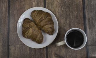 Two croissants are placed on a white plate, accompanied by a cup of coffee on a wooden table.