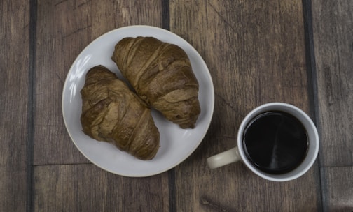 Two croissants are placed on a white plate, accompanied by a cup of coffee on a wooden table.