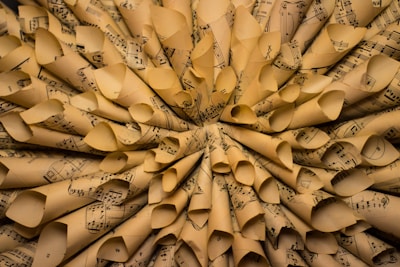 A close-up view of numerous rolled sheets of music paper arranged in a circular pattern, creating a textured and layered appearance. The paper is aged, with a yellowish tint and visible music notes and staffs printed on each sheet.