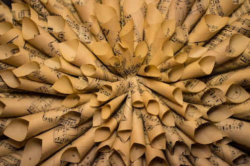 A close-up view of numerous rolled sheets of music paper arranged in a circular pattern, creating a textured and layered appearance. The paper is aged, with a yellowish tint and visible music notes and staffs printed on each sheet.