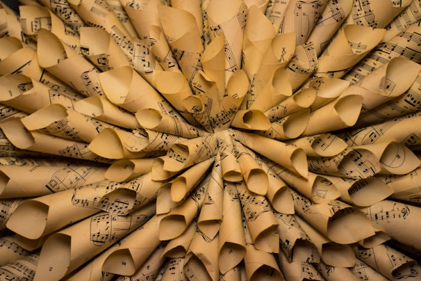 A close-up view of numerous rolled sheets of music paper arranged in a circular pattern, creating a textured and layered appearance. The paper is aged, with a yellowish tint and visible music notes and staffs printed on each sheet.