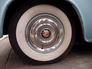 A close-up view of a vintage car wheel with a shiny chrome hubcap featuring a red and gold emblem in the center. The tire has a whitewall design, and the car body is painted light blue with some visible wear around the edges.