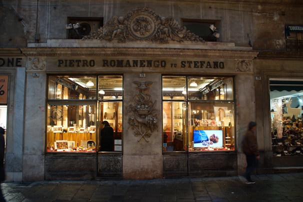 A historic storefront with elegant architectural details houses an interior filled with beautifully displayed products. The windows are warmly lit, showcasing various confectionery items inside. A person is visible inside admiring the displays, while another person walks by outside. The signage above the store reads 'Pietro Romanengo fu Stefano.'