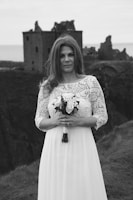 A woman in a white lace dress holds a bouquet while standing in front of the ruins of a castle on a grassy cliff. The surroundings suggest a moody, overcast day with a historic and romantic atmosphere.