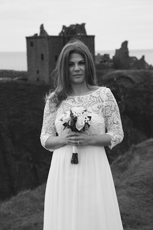 A woman in a white lace dress holds a bouquet while standing in front of the ruins of a castle on a grassy cliff. The surroundings suggest a moody, overcast day with a historic and romantic atmosphere.