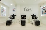 A modern nail salon interior featuring three manicure stations with black chairs and white cabinetry. The salon has bright lighting and is decorated in a minimalist style with a predominantly white color palette. Nail polish displays with a wide variety of colors line the walls, and the salon's logo is prominently displayed above the stations.