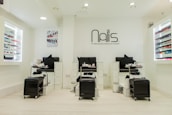 A modern nail salon interior featuring three manicure stations with black chairs and white cabinetry. The salon has bright lighting and is decorated in a minimalist style with a predominantly white color palette. Nail polish displays with a wide variety of colors line the walls, and the salon's logo is prominently displayed above the stations.
