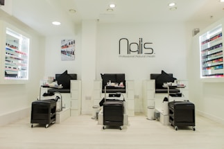 A modern nail salon interior featuring three manicure stations with black chairs and white cabinetry. The salon has bright lighting and is decorated in a minimalist style with a predominantly white color palette. Nail polish displays with a wide variety of colors line the walls, and the salon's logo is prominently displayed above the stations.