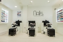 A modern nail salon interior featuring three manicure stations with black chairs and white cabinetry. The salon has bright lighting and is decorated in a minimalist style with a predominantly white color palette. Nail polish displays with a wide variety of colors line the walls, and the salon's logo is prominently displayed above the stations.