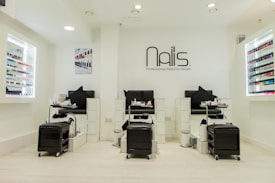 A modern nail salon interior featuring three manicure stations with black chairs and white cabinetry. The salon has bright lighting and is decorated in a minimalist style with a predominantly white color palette. Nail polish displays with a wide variety of colors line the walls, and the salon's logo is prominently displayed above the stations.