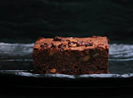 An artfully layered pistachio brownie with creamy frosting and crushed nuts, set against a chocolate brown background.