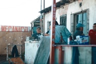 A group of people is gathered outside a mechanic shop. The setting appears informal, with individuals engaged in conversation or work-related activities. There are objects like a jack stand and various tools on a makeshift counter. The building is worn, with peeling paint and barred windows.