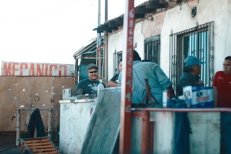 A group of people is gathered outside a mechanic shop. The setting appears informal, with individuals engaged in conversation or work-related activities. There are objects like a jack stand and various tools on a makeshift counter. The building is worn, with peeling paint and barred windows.