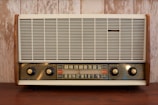A vintage radio with a wooden frame, featuring a large front grille and four control knobs. The tuning scale in the center displays frequency numbers.