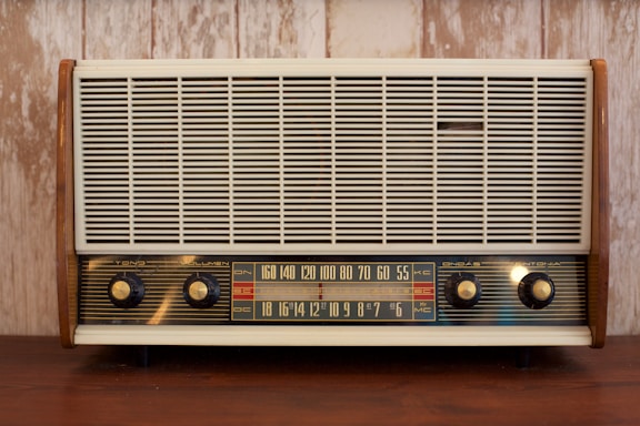 A vintage radio with a wooden frame, featuring a large front grille and four control knobs. The tuning scale in the center displays frequency numbers.