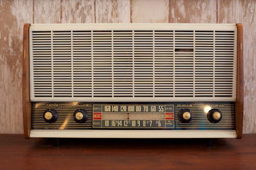 A vintage radio with a wooden frame, featuring a large front grille and four control knobs. The tuning scale in the center displays frequency numbers.