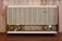 A vintage radio with a wooden frame, featuring a large front grille and four control knobs. The tuning scale in the center displays frequency numbers.