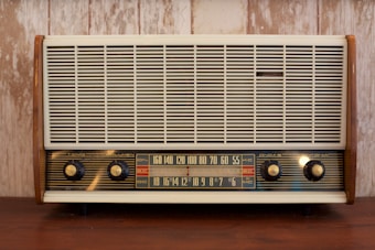 A vintage radio with a wooden frame, featuring a large front grille and four control knobs. The tuning scale in the center displays frequency numbers.