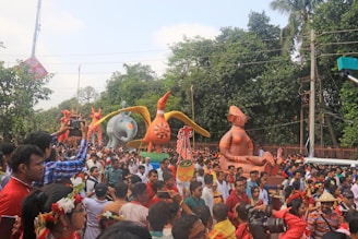 A large crowd of people participates in a colorful parade featuring giant, vibrant sculptures of animals and human figures. The scene is lively and festive, with many attendees wearing traditional attire and flower crowns. There are lush green trees in the background, and the sky is partly cloudy.