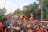 A large crowd of people participates in a colorful parade featuring giant, vibrant sculptures of animals and human figures. The scene is lively and festive, with many attendees wearing traditional attire and flower crowns. There are lush green trees in the background, and the sky is partly cloudy.