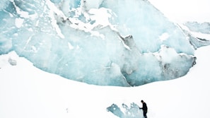 A person stands at the foot of a massive glacier wall, surrounded by vast expanses of snow and ice. The glacier displays varying shades of blue and white, with dark gray crevices adding texture. The scene is serene and remote, highlighting the scale of the natural ice formation in comparison to the solitary figure.