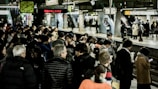 A busy train station platform filled with people waiting, some engaging with their phones, others chatting. Bright lights illuminate the area, with signs and a clock overhead indicating train times. The platform is bustling with activity, showcasing people in coats, jackets, and diverse attire.