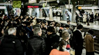 A busy train station platform filled with people waiting, some engaging with their phones, others chatting. Bright lights illuminate the area, with signs and a clock overhead indicating train times. The platform is bustling with activity, showcasing people in coats, jackets, and diverse attire.
