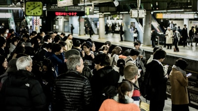 A busy train station platform filled with people waiting, some engaging with their phones, others chatting. Bright lights illuminate the area, with signs and a clock overhead indicating train times. The platform is bustling with activity, showcasing people in coats, jackets, and diverse attire.