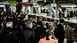A busy train station platform filled with people waiting, some engaging with their phones, others chatting. Bright lights illuminate the area, with signs and a clock overhead indicating train times. The platform is bustling with activity, showcasing people in coats, jackets, and diverse attire.