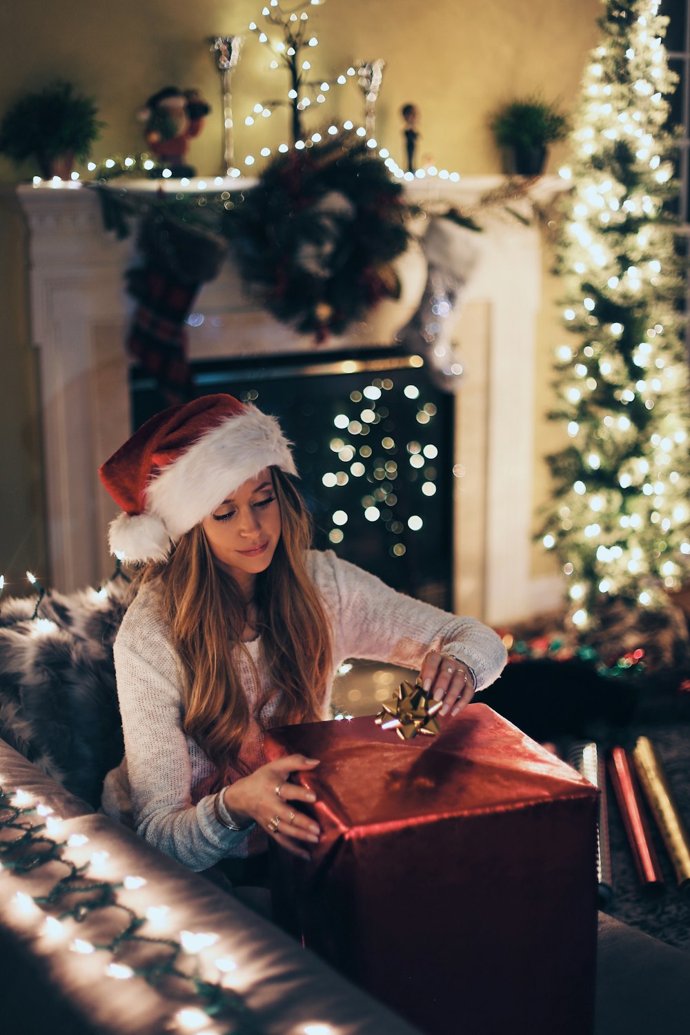 woman putting ribbon in red gift box near lighted Christmas tree inside room