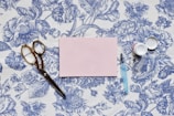 A vintage-style wallpaper with intricate blue floral designs serves as the background for a flat lay arrangement. A pair of gold scissors, a light blue pen with a transparent cap, and an open small container are placed beside a pink rectangular card.