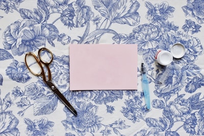 A vintage-style wallpaper with intricate blue floral designs serves as the background for a flat lay arrangement. A pair of gold scissors, a light blue pen with a transparent cap, and an open small container are placed beside a pink rectangular card.