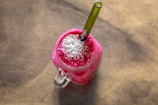 A bright pink smoothie topped with shredded coconut is served in a clear glass jar on a wooden surface. A green straw is inserted into the drink, and there are visible bubbles on the surface.