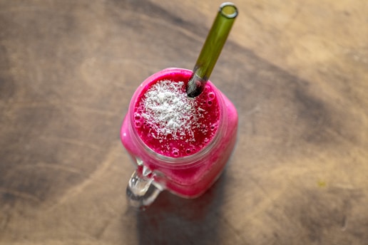 A bright pink smoothie topped with shredded coconut is served in a clear glass jar on a wooden surface. A green straw is inserted into the drink, and there are visible bubbles on the surface.