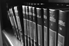 A row of hardcover books lined up on a shelf, featuring prominently displayed titles and authors on their spines. The image is captured in black and white, giving it a classic and timeless feel.