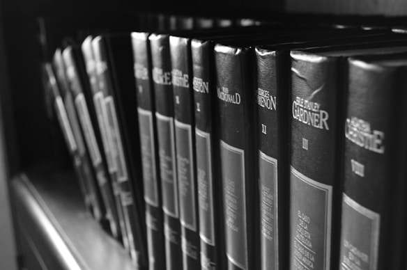 A row of hardcover books lined up on a shelf, featuring prominently displayed titles and authors on their spines. The image is captured in black and white, giving it a classic and timeless feel.