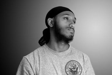 A black and white portrait of a person wearing a durag and a T-shirt with a high school emblem. The person is looking slightly upward with a thoughtful expression. The background is a gradient from dark to light gray.